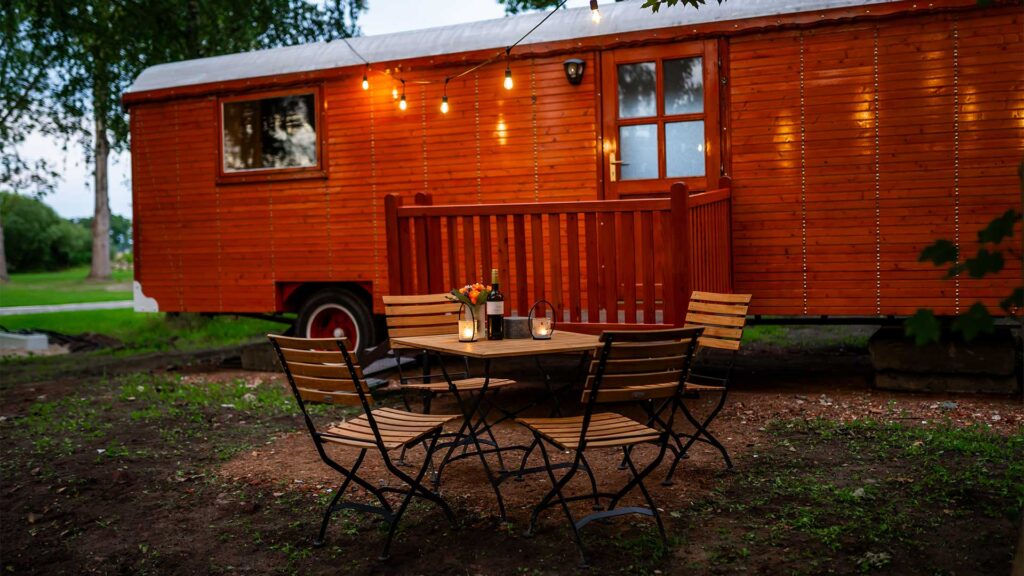 Ein Foto eines der Campingwagen des Schlosses. Davor steht in der Abenddämmerung ein Holztisch mit Stühlen und einer Lichterkette darüber.